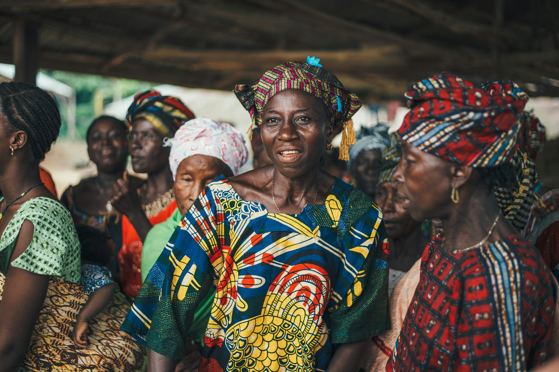 A group of women in brightly patterned dresses and headwraps stand together under a village shelter, one of them smiling towards the camera.