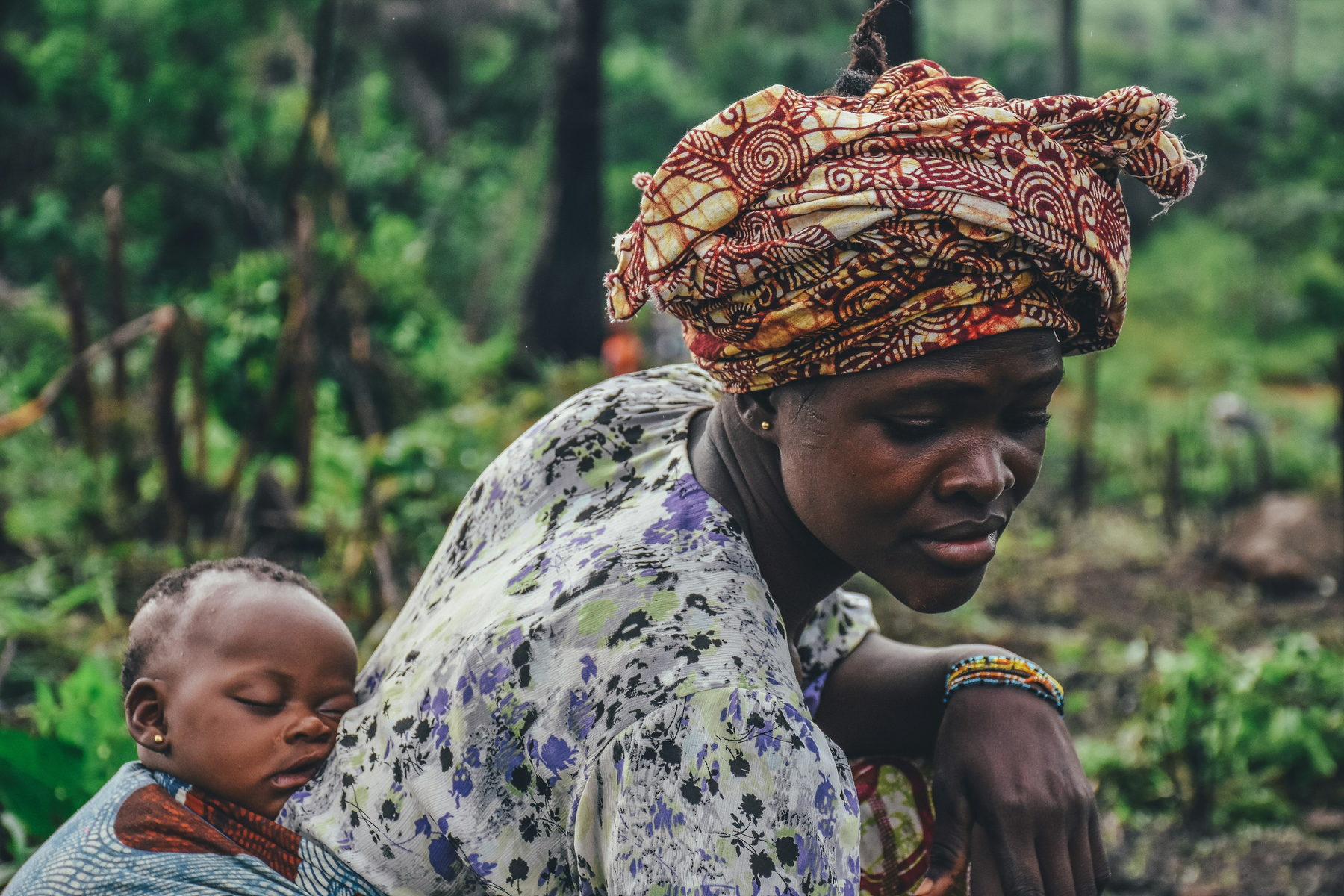 A woman in a patterned headwrap works in a cassava field with her sleeping baby carried in a wrap on her back.